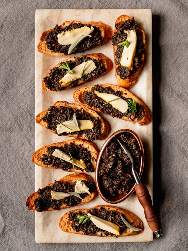 An overhead shot shows a platter of vegan mushroom duxelles crostini with little wedges of vegan cheese on top of each crostini; all garnished with small leaves of fresh herbs.