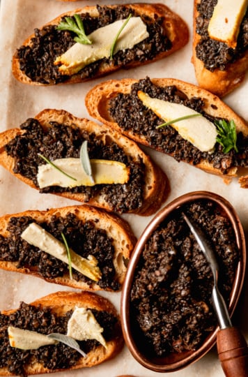 An up close, overhead shot shows a platter of vegan mushroom duxelles crostini with little wedges of vegan cheese on top of each crostini; all garnished with small leaves of fresh herbs.