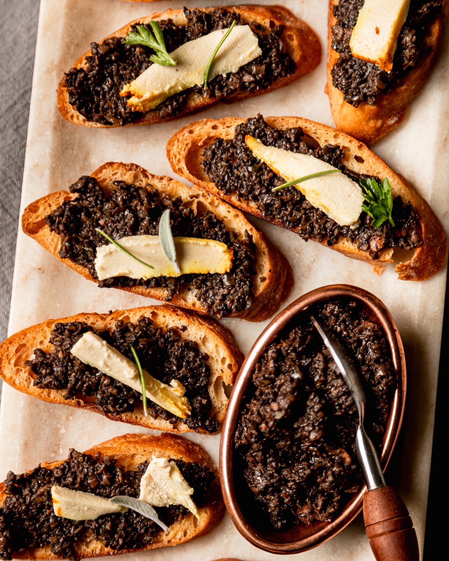 An up close, overhead shot shows a platter of vegan mushroom duxelles crostini with little wedges of vegan cheese on top of each crostini; all garnished with small leaves of fresh herbs.
