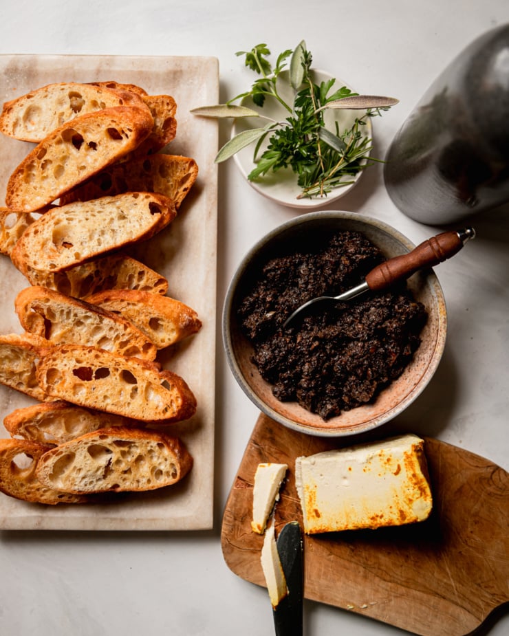 An overhead shot shows toasted rostini on a plate, a small. bowl of herbs, a bottle of olive oil, a bowl of mushroom duxelles, and a wedge of vegan cheese on a small wooden cutting board.