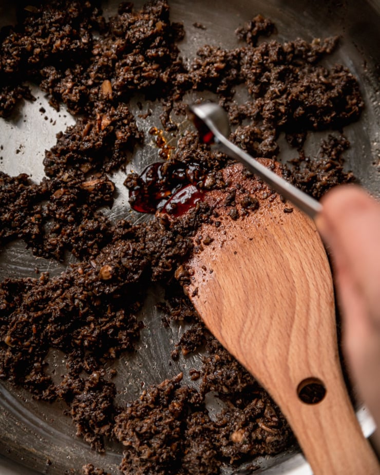 An up close, overhead shot shows Tamari being added by the 1/2 teaspoon to a skillet filled with vegan mushroom duxelles.