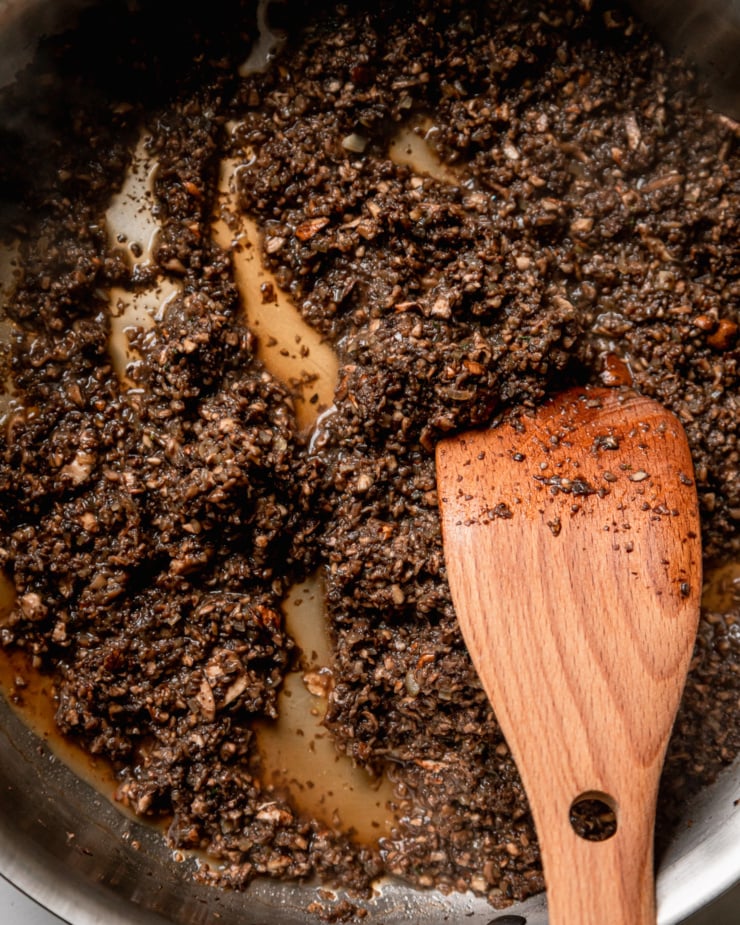 An up close, overhead shot shows finely minced mushrooms and shallots letting off liquid in a skillet with a wooden spoon.