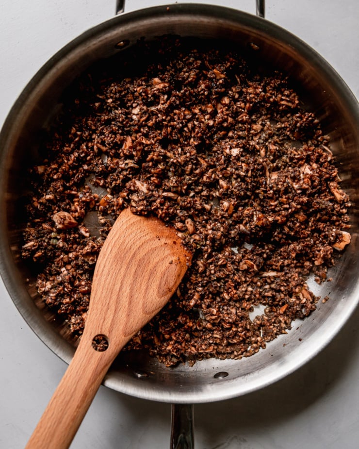 An overhead shot shows the beginning cooking phase of vegan mushroom duxelles in a stainless steel skillet with a wooden spoon sticking out.