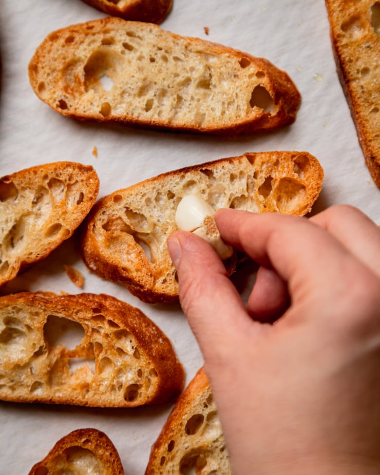 An up close, overhead shot shows a hand rubbing a halved garlic clove into toasted slices of baguette.