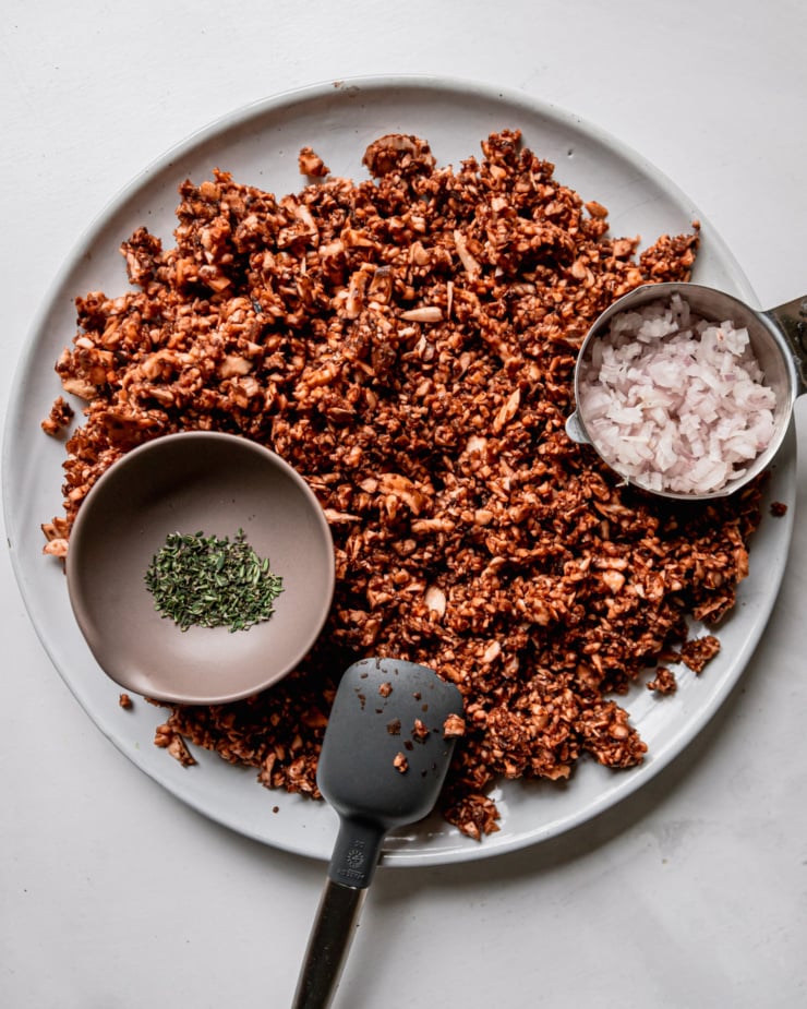 An overhead shot shows a plate filled with finely chopped mushrooms and one small bowl of minced fresh thyme, and a measuring cup of finely minced shallot. A spatula is also hanging off of the plate.