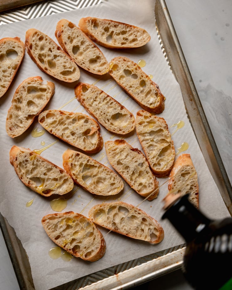 An overhead shot shows olive oil being drizzled over baguette slices on a parchment-lined baking sheet.