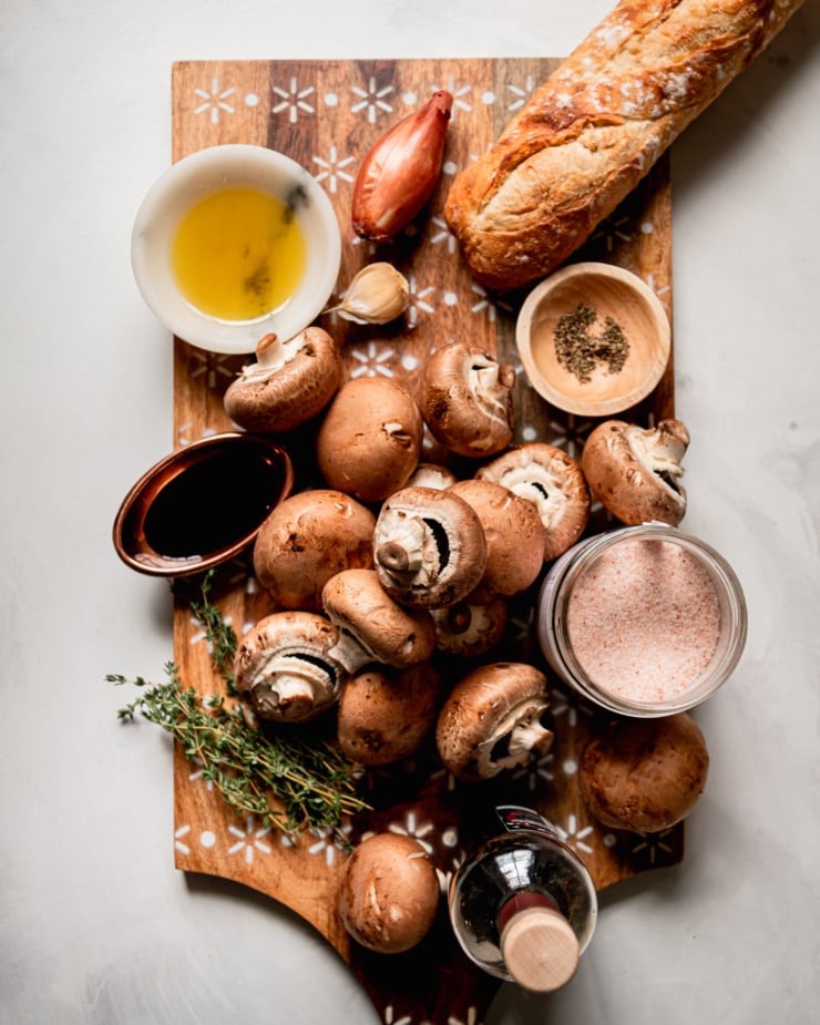 An overhead shot shows ingredients for a vegan appetizer: baguette, mushrooms, olive oil, salt, pepper, thyme, shallot, garlic, balsamic vinegar, thyme, and Tamari; all on a wooden board.