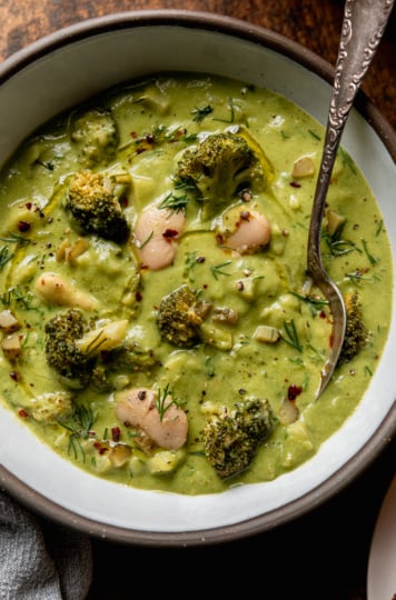An up close overhead shot shows a bowl of broccoli dill pickle soup with white beans and chopped potatoes. A spoon is sticking out of the bowl. The soup is garnished with chili flakes, chopped fresh dill, and swirls of olive oil.