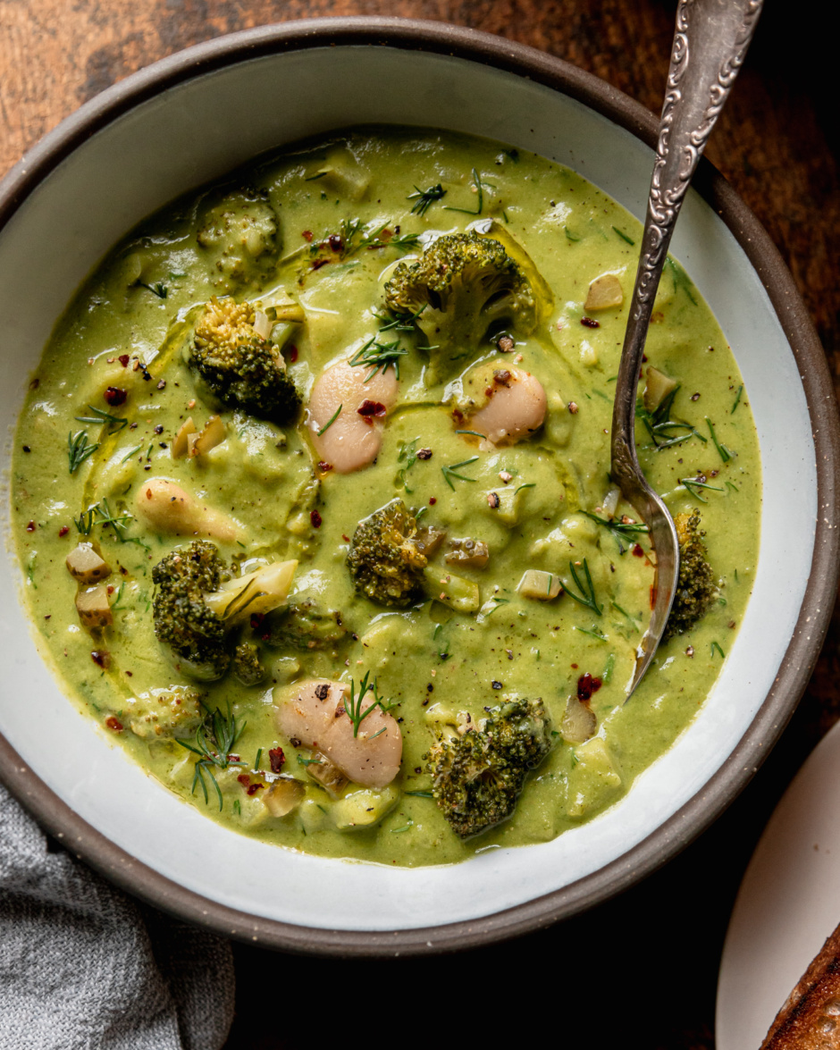 An up close overhead shot shows a bowl of broccoli dill pickle soup with white beans and chopped potatoes. A spoon is sticking out of the bowl. The soup is garnished with chili flakes, chopped fresh dill, and swirls of olive oil.