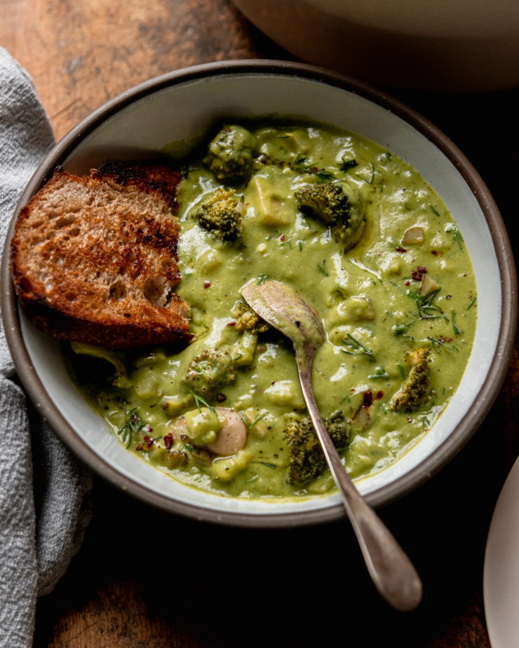 A 3/4 angle shot shows a bowl of broccoli dill pickle soup with a toasted piece of sourdough bread and a spoon sticking out.