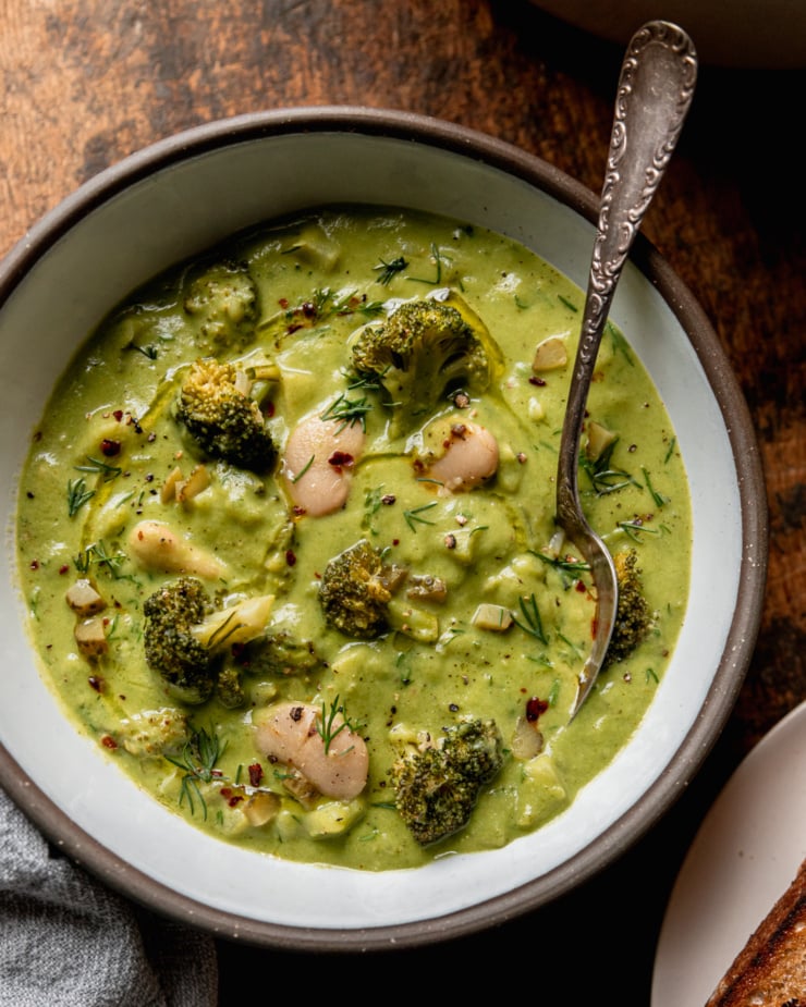 An up close overhead shot shows a bowl of broccoli dill pickle soup with white beans and chopped potatoes. A spoon is sticking out of the bowl. The soup is garnished with chili flakes, chopped fresh dill, and swirls of olive oil.