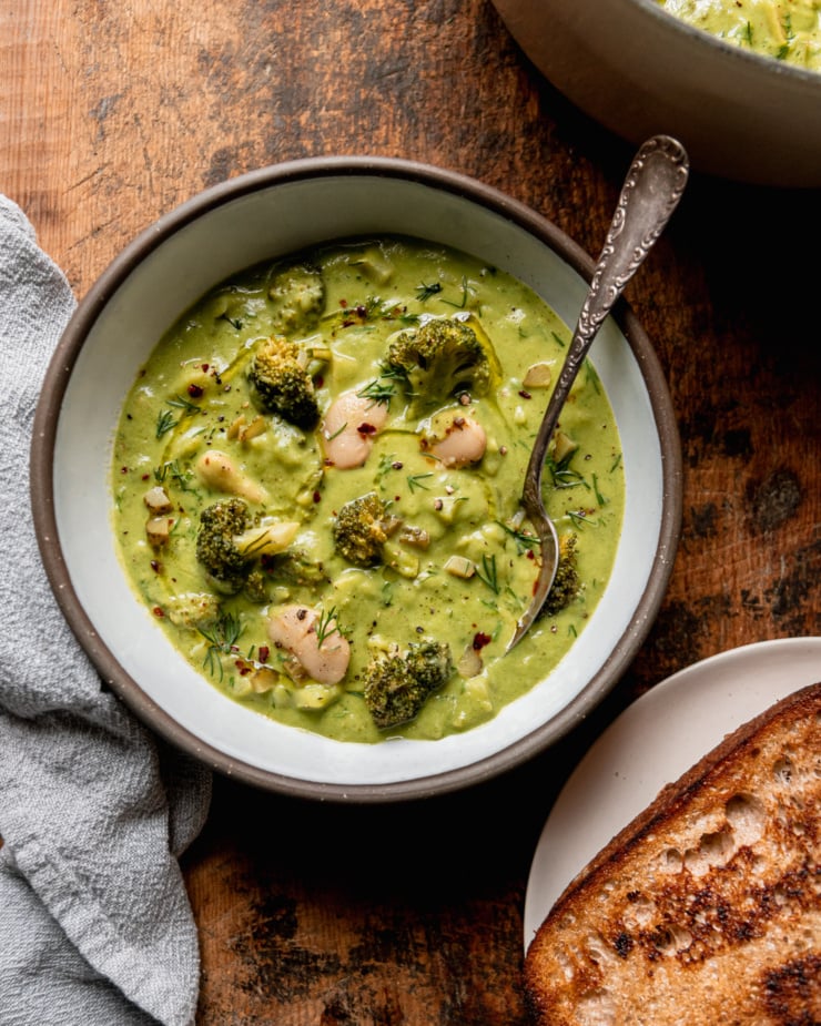 An overhead shot shows a bowl of broccoli dill pickle soup with white beans and chopped potatoes. A spoon is sticking out of the bowl. The soup is garnished with chili flakes, chopped fresh dill, and swirls of olive oil. A plate with toasted sourdough bread is nearby.
