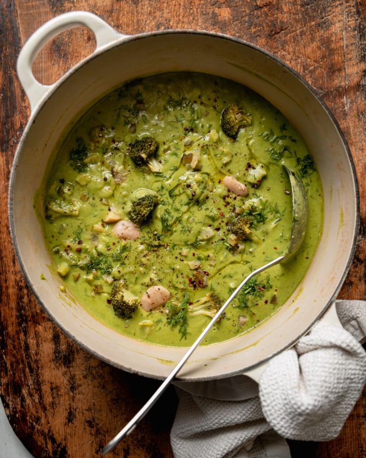 An overhead shot shows a pot of vegan broccoli dill pickle soup with butter beans and potatoes. The top of the soup is garnished with chili flakes, swirls of olive oil, and chopped fresh dill. A ladle is sticking out of the pot.