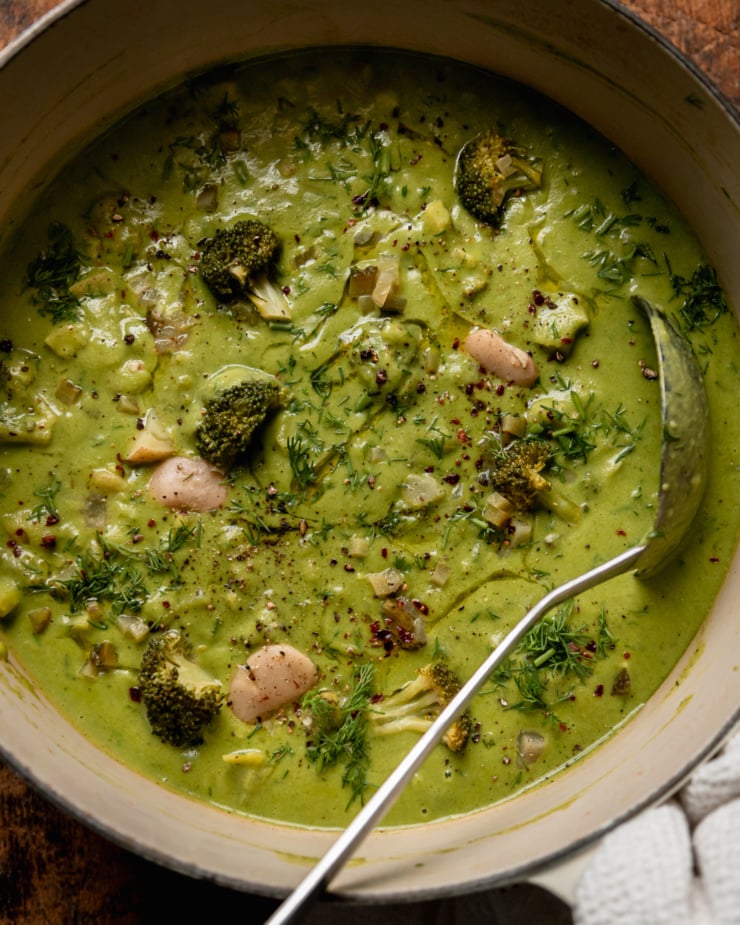 An up close overhead shot shows a pot of vegan broccoli dill pickle soup with butter beans and potatoes. The top of the soup is garnished with chili flakes, swirls of olive oil, and chopped fresh dill. A ladle is sticking out of the pot.