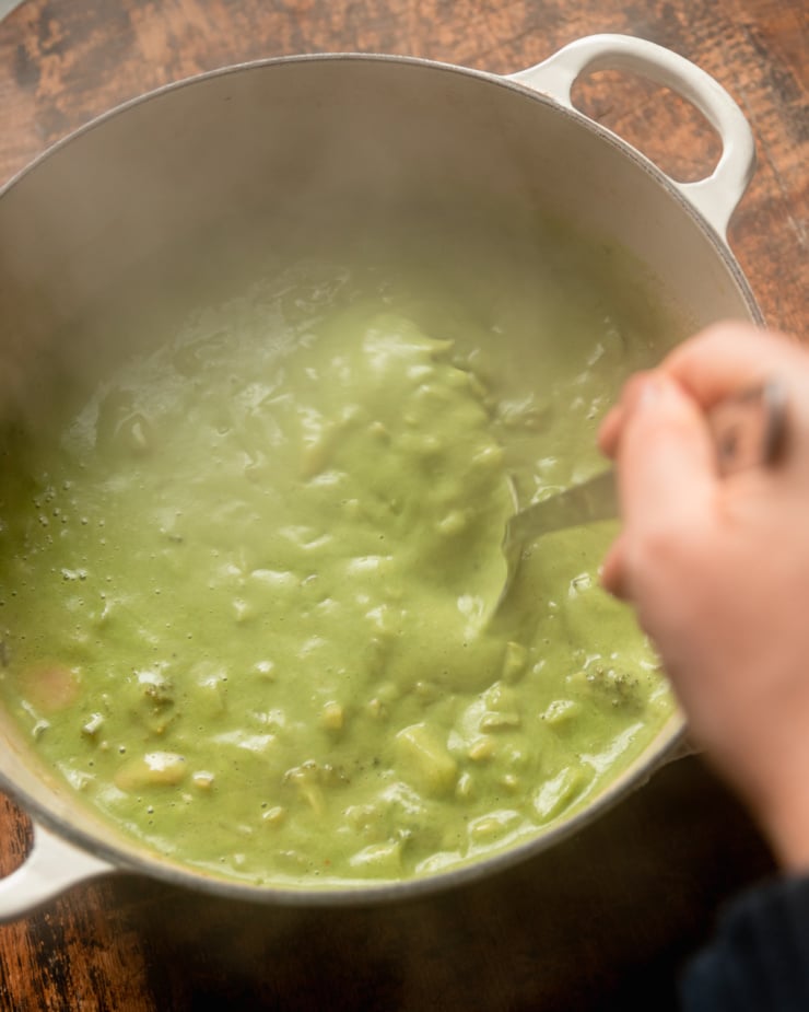 An overhead shot shows a hand using a ladle to stir up a steamy and creamy light green pot of soup.