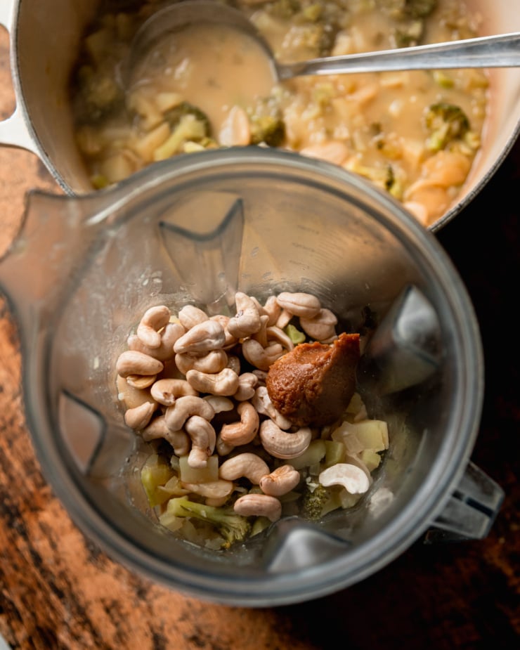 An overhead shot shows a blender filled with some hot soup and soaked cashews and miso on top. The pot of soup is in the background.
