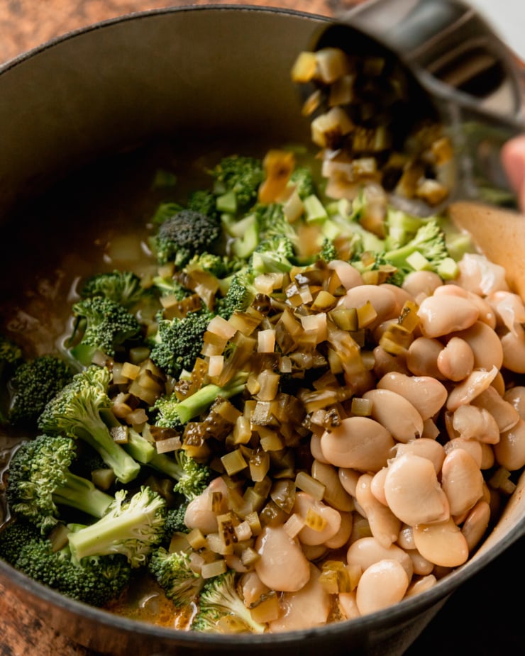 An overhead shot shows chopped dill pickles being added to a pot with butter beans, broccoli, potatoes, onions, celery, and vegetable stock.