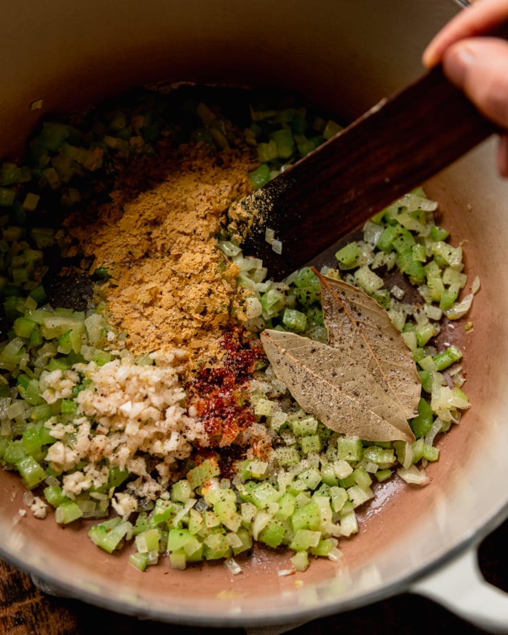 An overhead shot shows sautéed onions and celery on a pot with spices and bay leaves added on top.