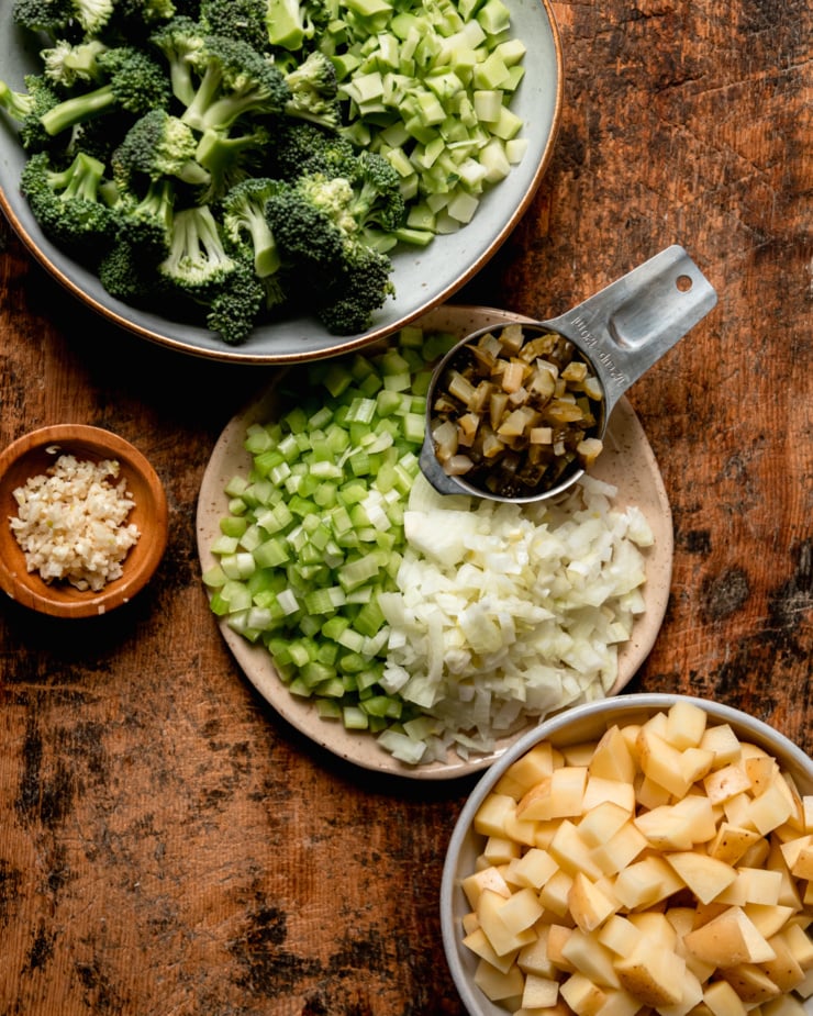 An overhead shot shows plates and bowls with chopped broccoli, celery, onions, dill pickles, garlic, and potatoes.