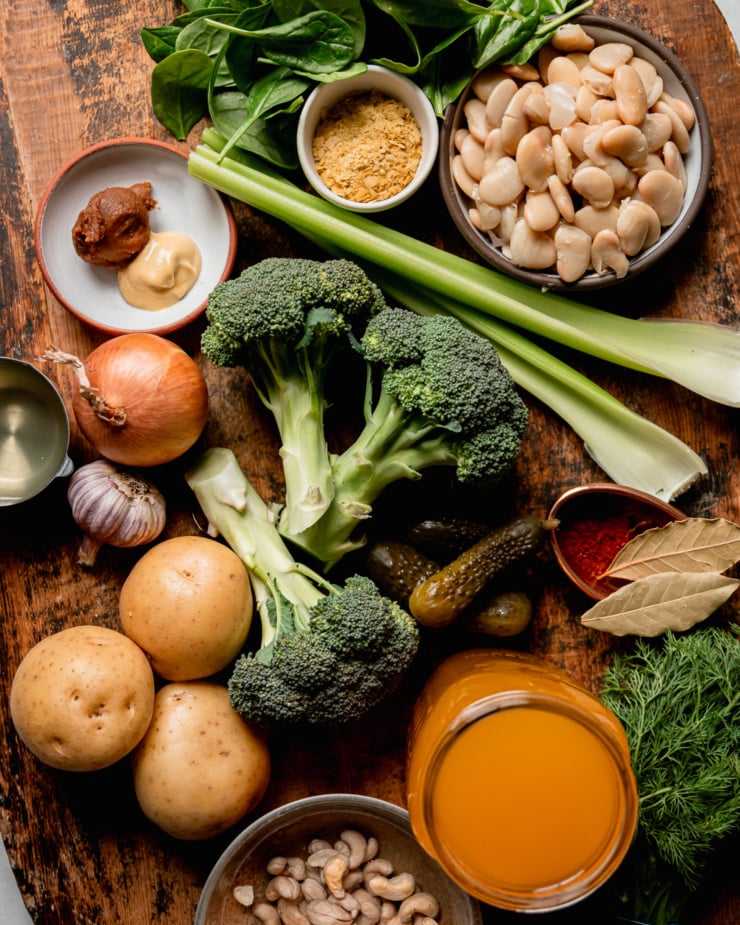 An overhead shot shows ingredients for a soup, featuring broccoli, potatoes, white butter beans, nutritional yeast, spices, vegetable stock, and more.