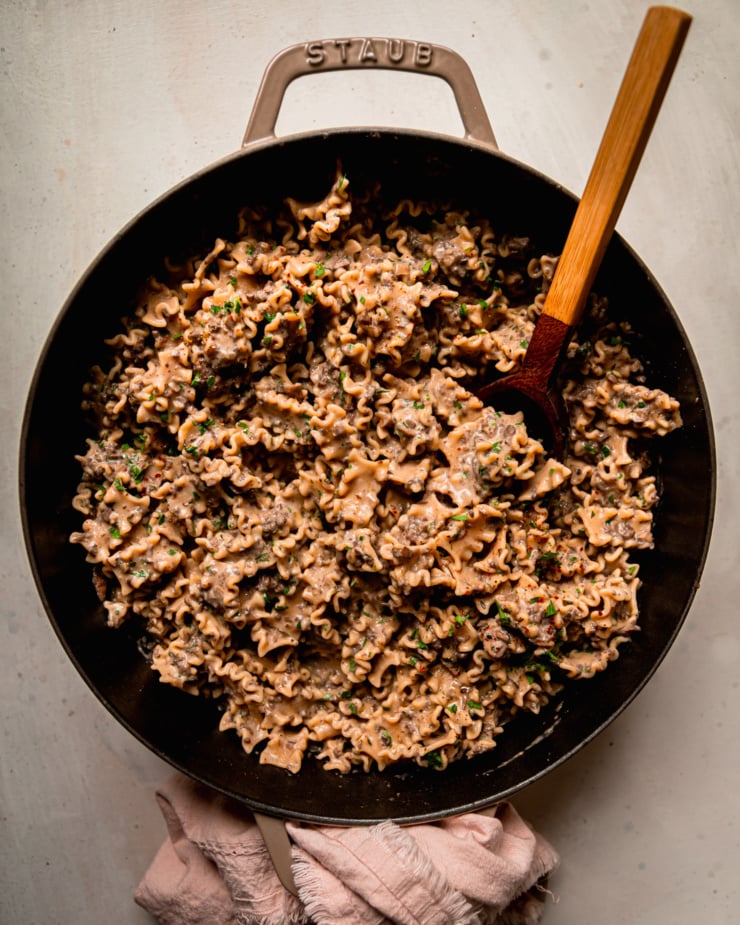An overhead shot shows a braiser pot filled with creamy vegan pasta dressed in a finely chopped mushroom and miso sauce. A wooden serving spoon is sticking out of the pot and a linen is wrapped around the handle on one side.