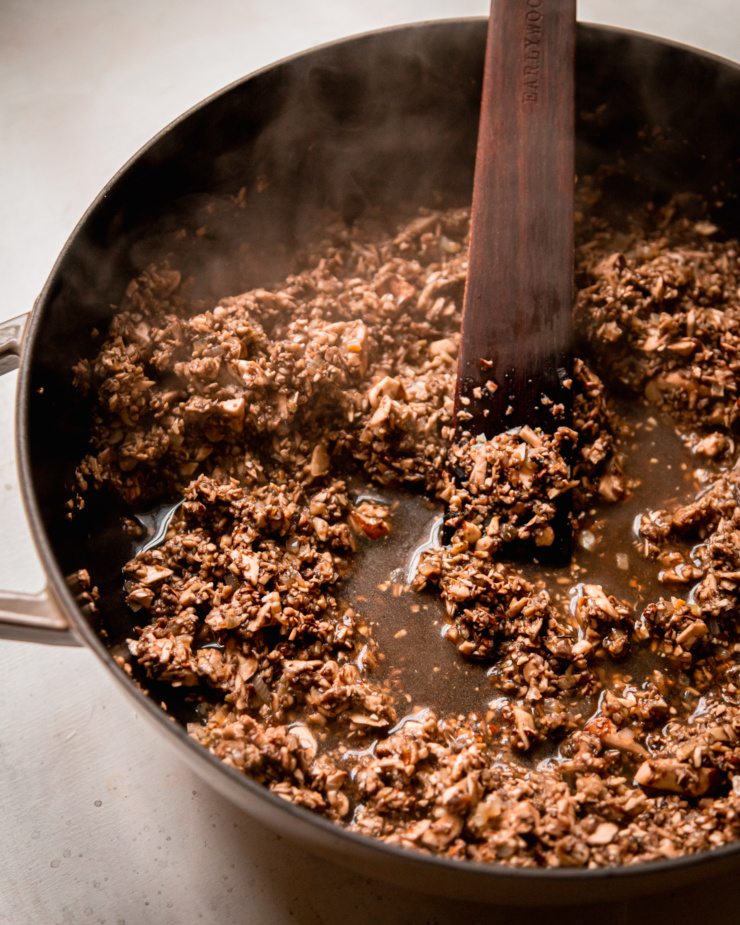 A 3/4 angle shot shows finely chopped mushrooms letting off a lot of liquid and steam in a braiser style pot.