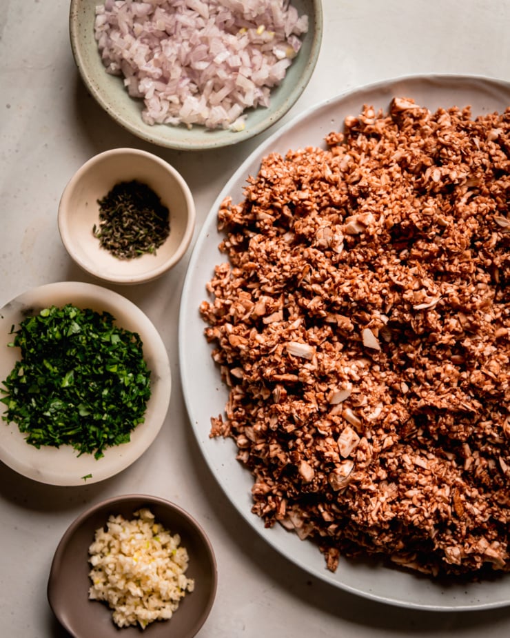 An overhead shot shows a bowl of small dice shallots, a plate of finely minced mushrooms, a bowl with minced garlic, a bowl with chopped parsley, and a small bowl with minced fresh thyme.