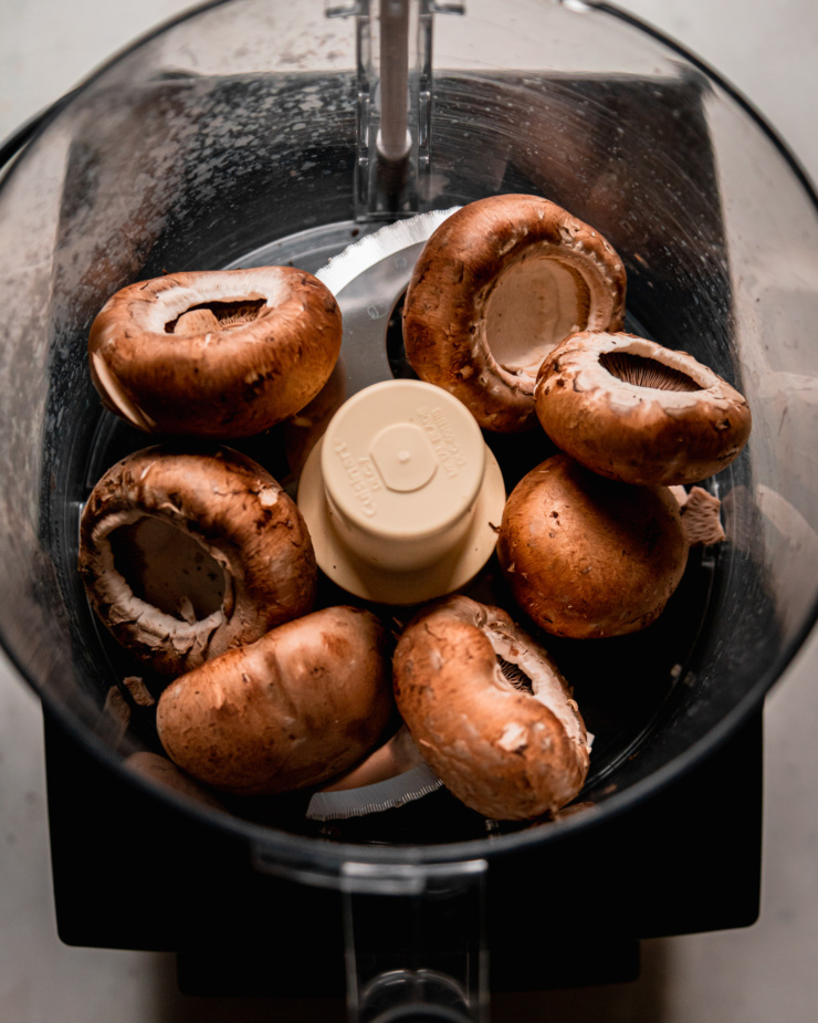 An overhead shot shows whole cremini mushroom caps in the bowl of a food processor.