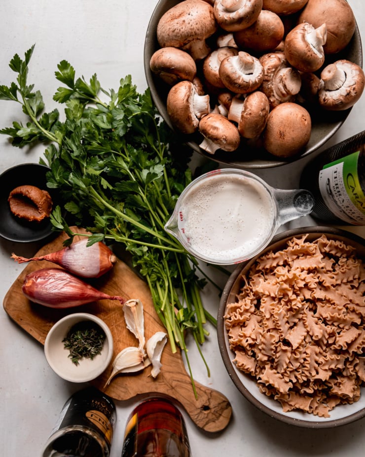An overhead shot shows ingredients used in a creamy vegan pasta recipe: cremini mushrooms, olive oil, non-dairy "creamer," sherry vinegar, Tamari, garlic, thyme, shallots, light miso, and parsley.