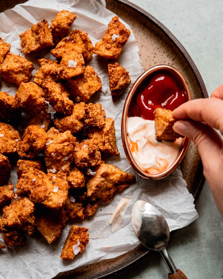 An overhead shot shows a plate filled with crispy air fryer tofu pieces. A hand is dipping a piece into a little bowl with spicy mayo and ketchup inside.