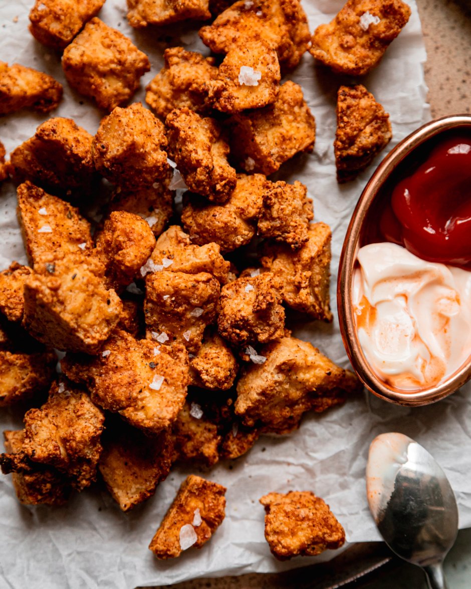 An up close, overhead shot shows crispy pieces of air fryer tofu on a plate lined with crinkled parchment paper. To the side is some spicy mayo and ketchup for dipping.