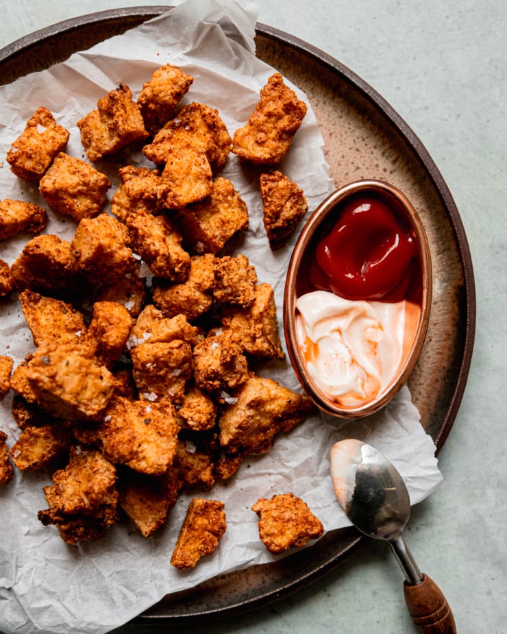 An overhead shot shows crispy pieces of air fryer tofu on a plate lined with crinkled parchment paper. To the side is some spicy mayo and ketchup for dipping.