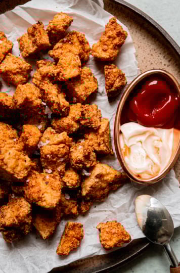 An overhead shot shows crispy pieces of air fryer tofu on a plate lined with crinkled parchment paper. To the side is some spicy mayo and ketchup for dipping.
