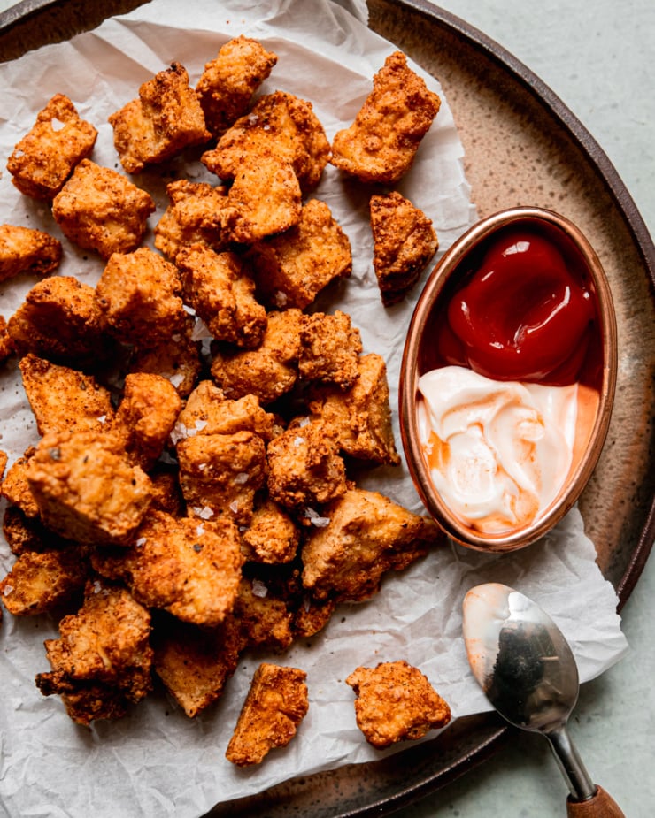 An overhead shot shows crispy pieces of air fryer tofu on a plate lined with crinkled parchment paper. To the side is some spicy mayo and ketchup for dipping.