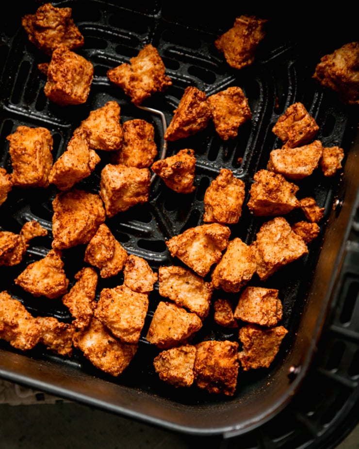 An up close, overhead shot shows jagged and crispy pieces of tofu that are thoroughly golden brown. The pieces are inside of an air fryer basket.