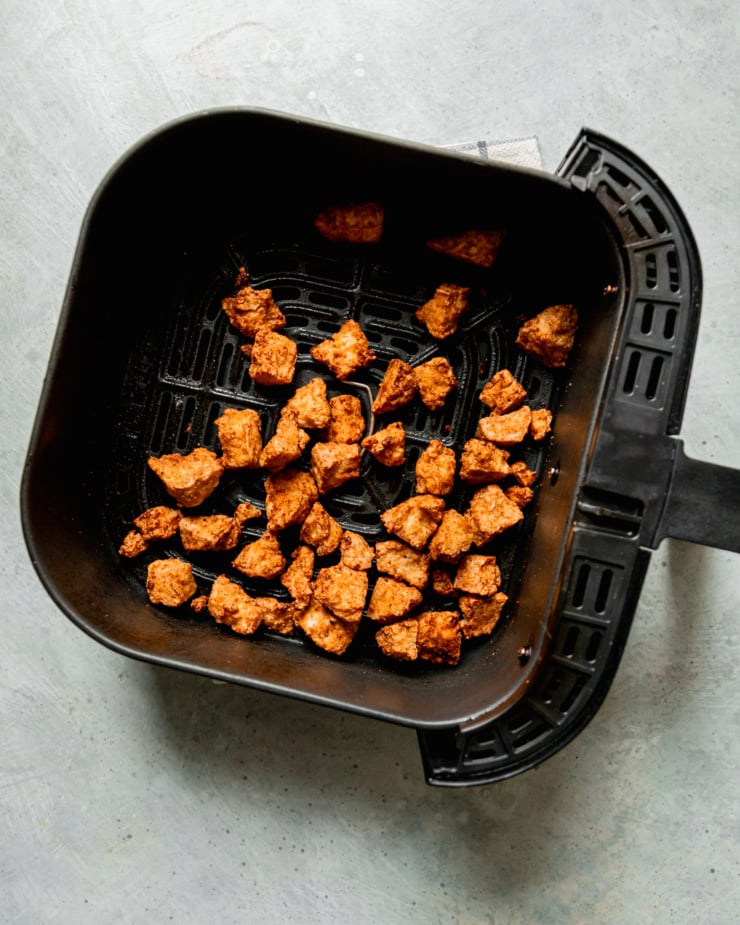 An overhead shot shows jagged and crispy pieces of tofu that are thoroughly golden brown. The pieces are inside of an air fryer basket.