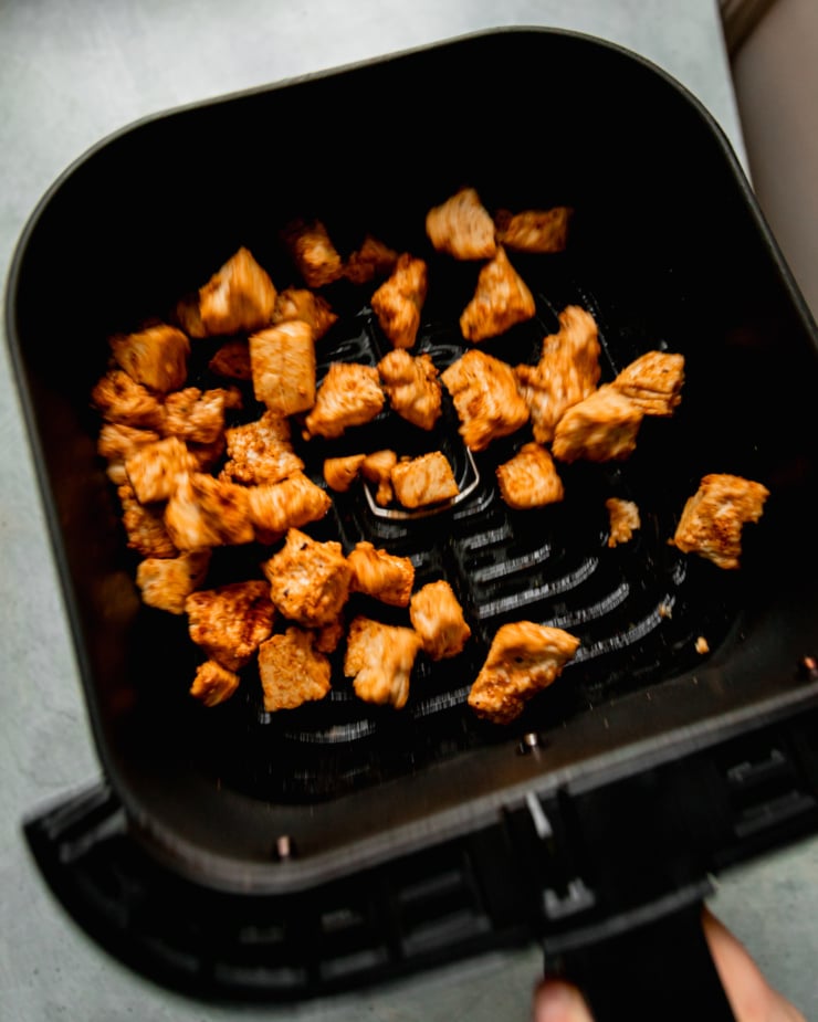 An overhead shot shows an air fryer basket being shaken with pieces of golden tofu inside.