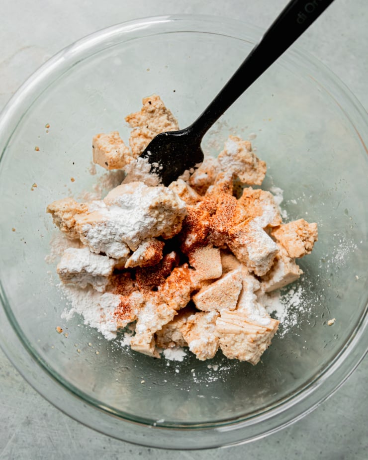 An overhead shot shows a bowl with tofu pieces, arrowroot starch, and spices. A spatula is sticking out of the bowl.
