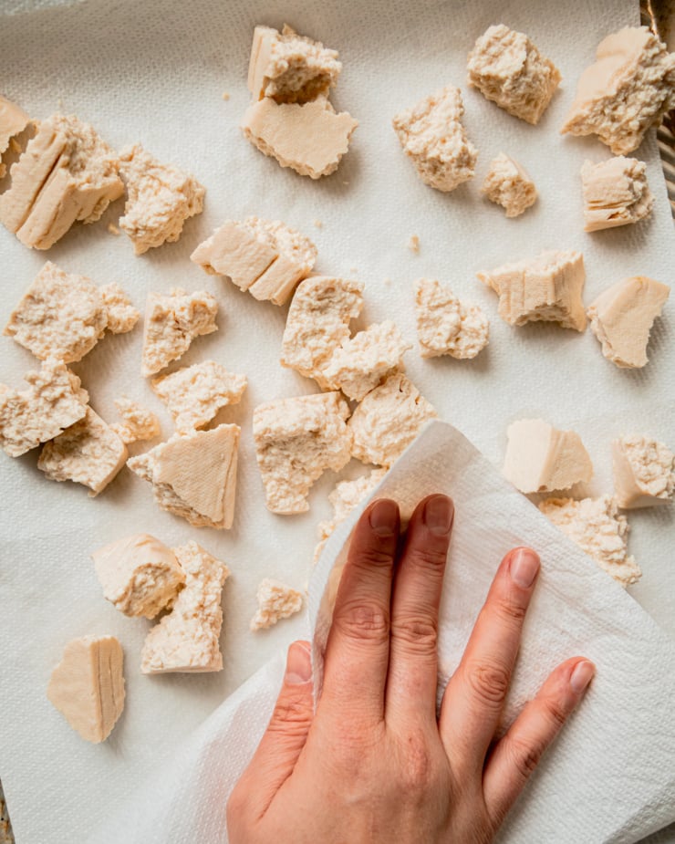 An overhead shot shows a hand using paper towel to blot torn pieces of tofu dry.
