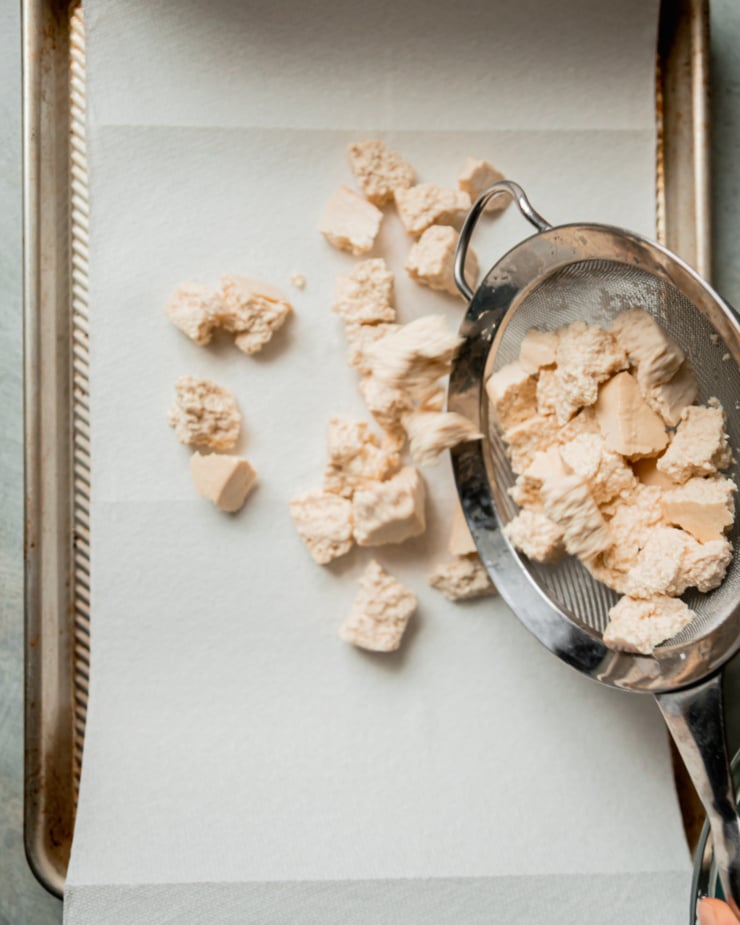 An overhead shot shows pieces of tofu in a fine mesh strainer being emptied out onto a baking sheet lined with paper towel.