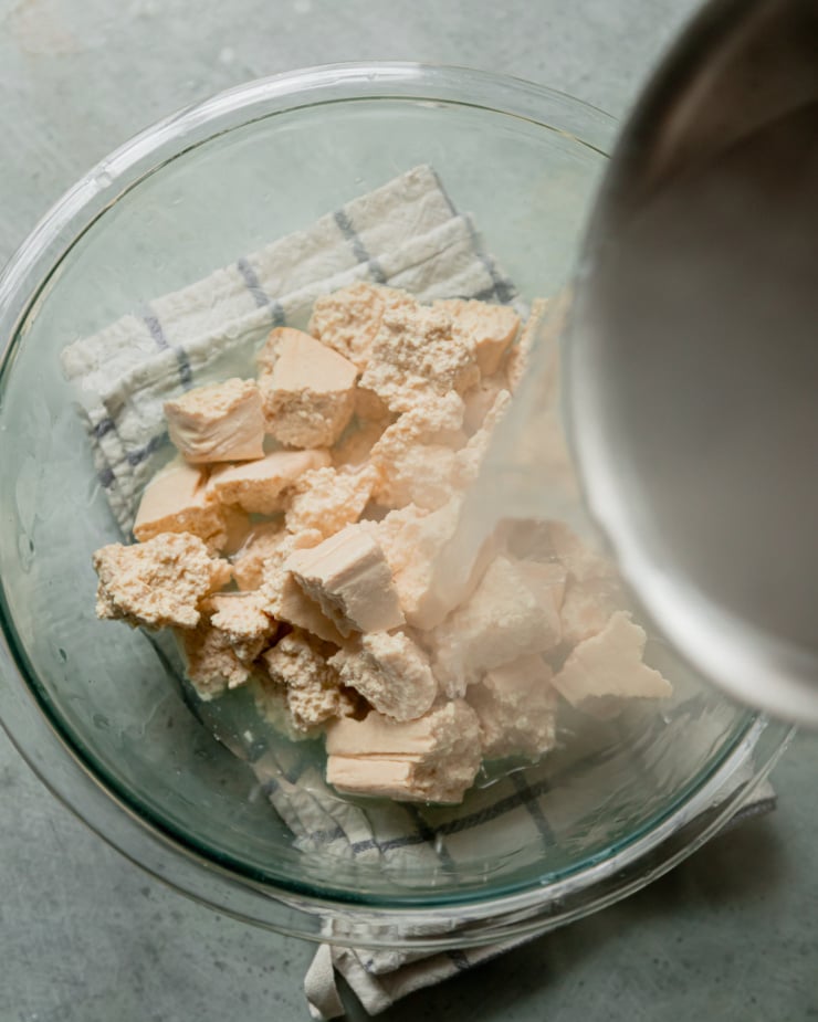 An overhead shot shows boiling salted water being poured over torn pieces of tofu in a bowl.