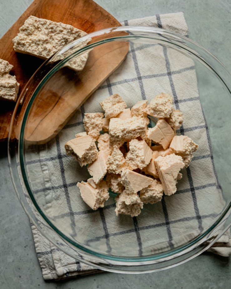 An overhead shot shows torn pieces of tofu in a bowl. The block of tofu is on a board to the side.