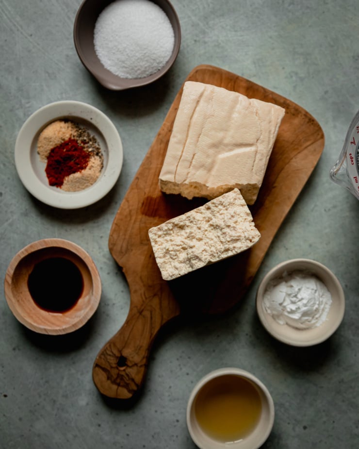 An overhead shot shows ingredients needed for crispy tofu in the air fryer: firm tofu, arrowroot starch, avocado oil, tamari, spices, and salt.
