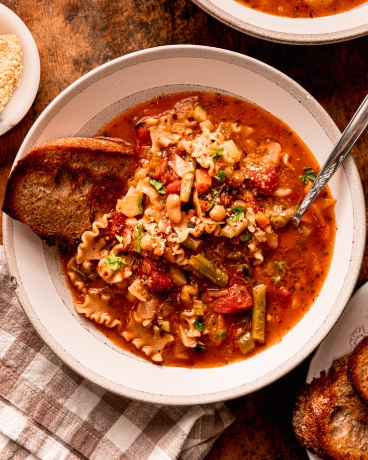 An overhead shot shows am individual bowl of vegan minestrone soup with a spoon sticking out. A toasted piece of sourdough bread is also nested into the bowl.