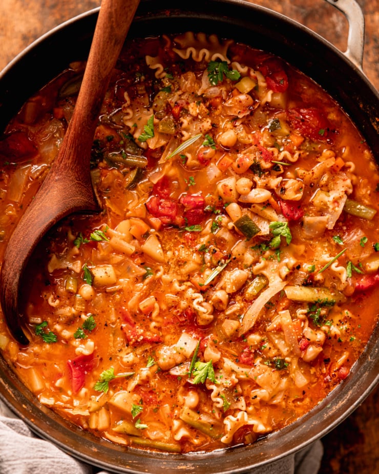 An up close, overhead shot shows a pot of vegan minestrone soup. A wooden ladle is sticking out of the pot.