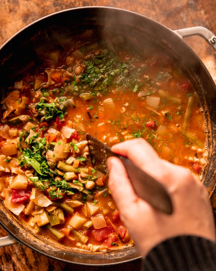 An overhead shows a hand using a wooden utensil to stir chopped parsley into a pot of soup.