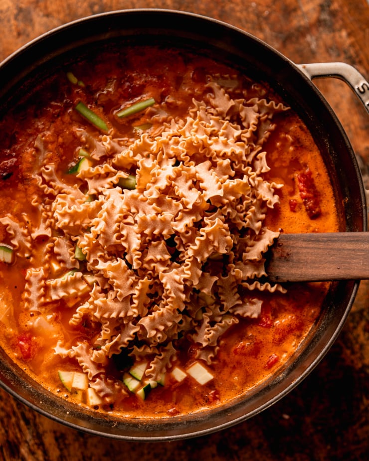 An overhead shot shows dry whole wheat pasta added to a pot of soup. A wooden utensil is sticking out of the pot. The pasta shape looks like tiny lasagna noodles.