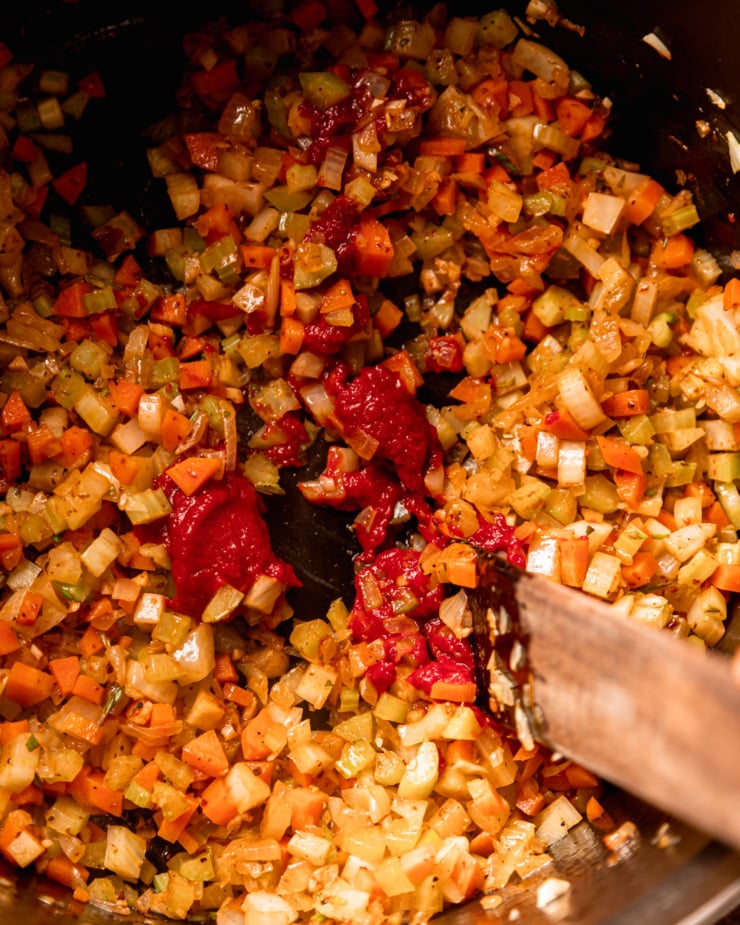 An overhead shot shows tomato paste being stirred into a mixture of sautéed vegetables in a pot.