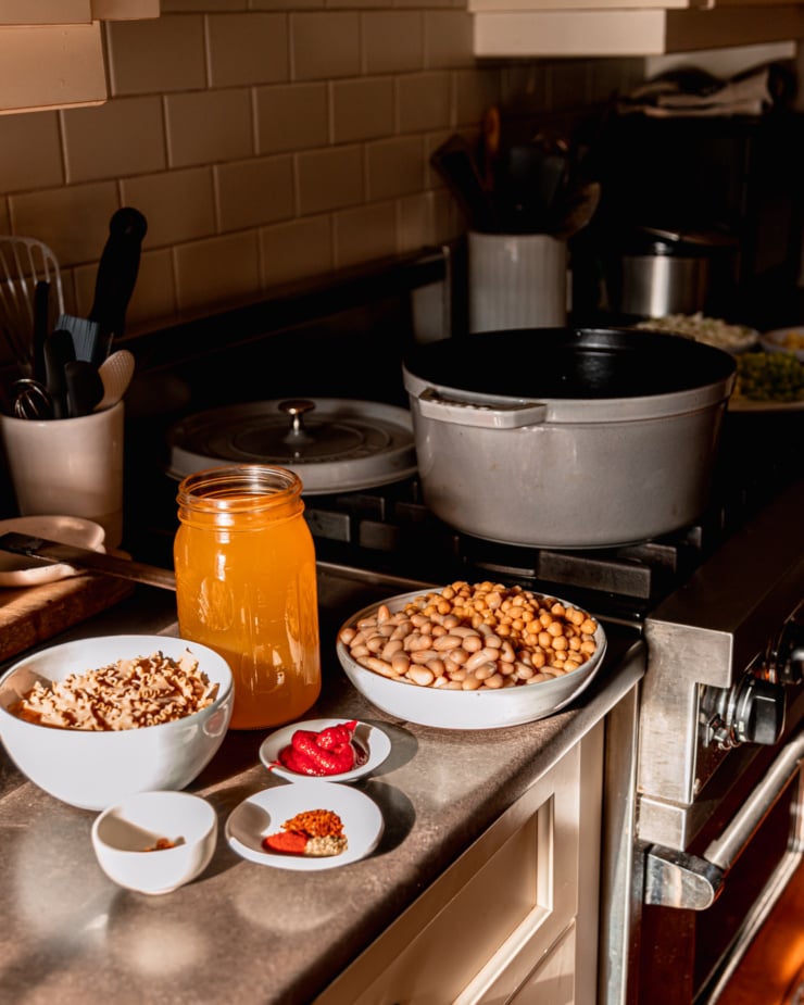 A slight 3/4 angle image shows a kitchen stove with a pot on top. Nearby on the counter is a bowl of mixed beans, a jar of vegetable stock, tomato paste, a bowl of measured out spices, and a bowl of dry pasta.