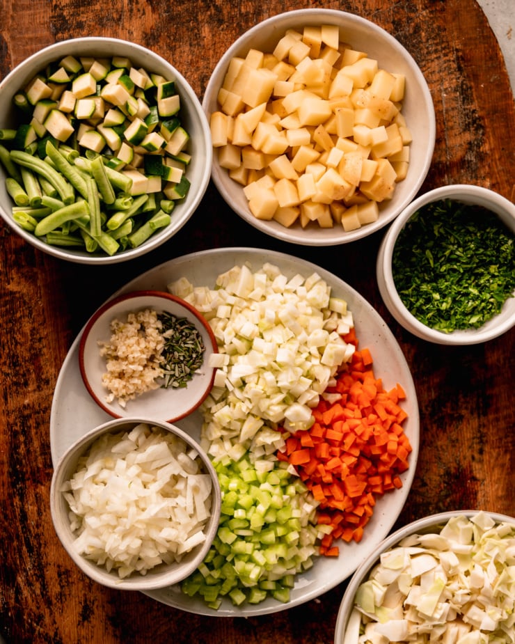 An overhead shot shows prepped and chopped green beans, zucchini, potatoes, parsley, cabbage, carrots, celery, fennel, onions, garlic, and rosemary.